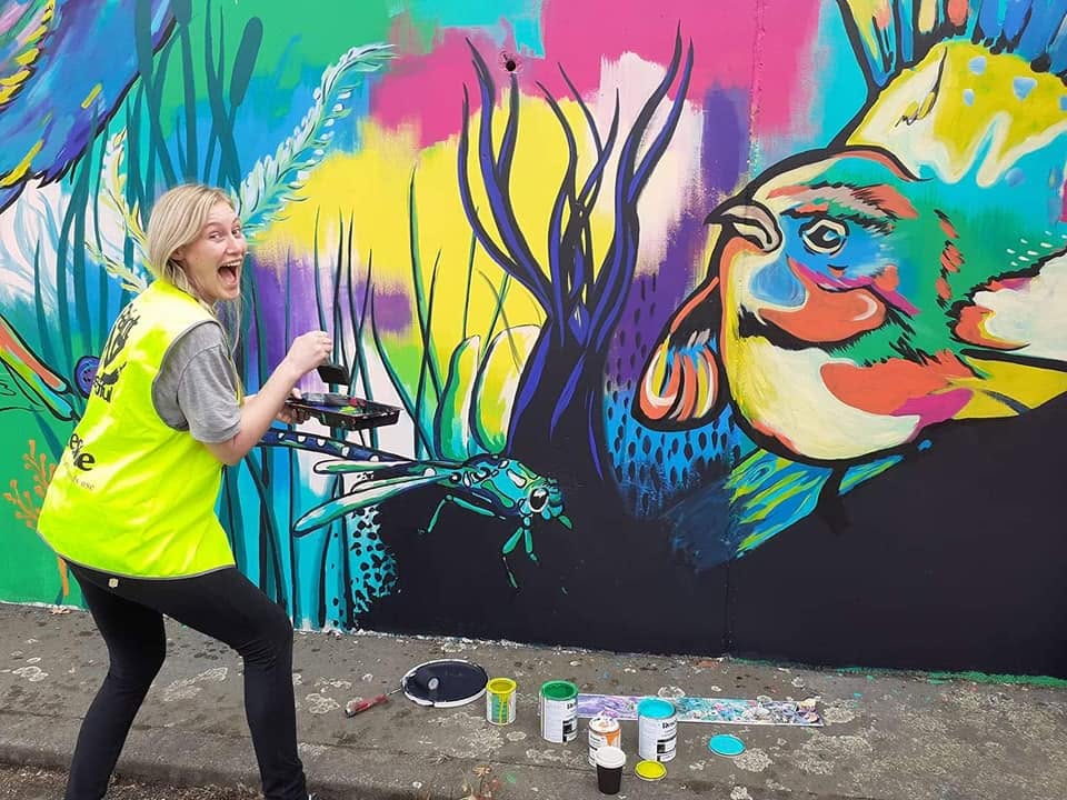 A muralist laughing while painting colourful native bird and insect details on a mural