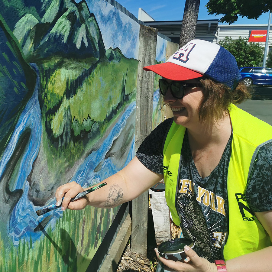 Laura painting a mountain and river landscape mural detail outdoors