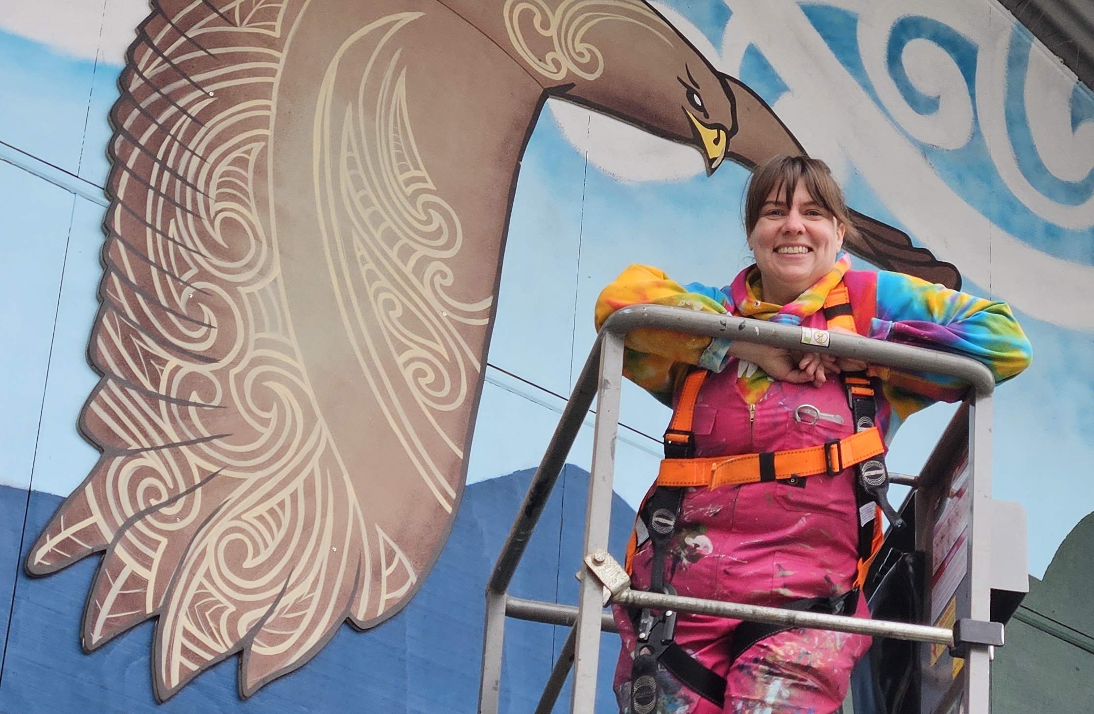 Mel Eaton on a lift platform in front of the In Our Hands mural, wearing a safety harness and paint-covered overalls