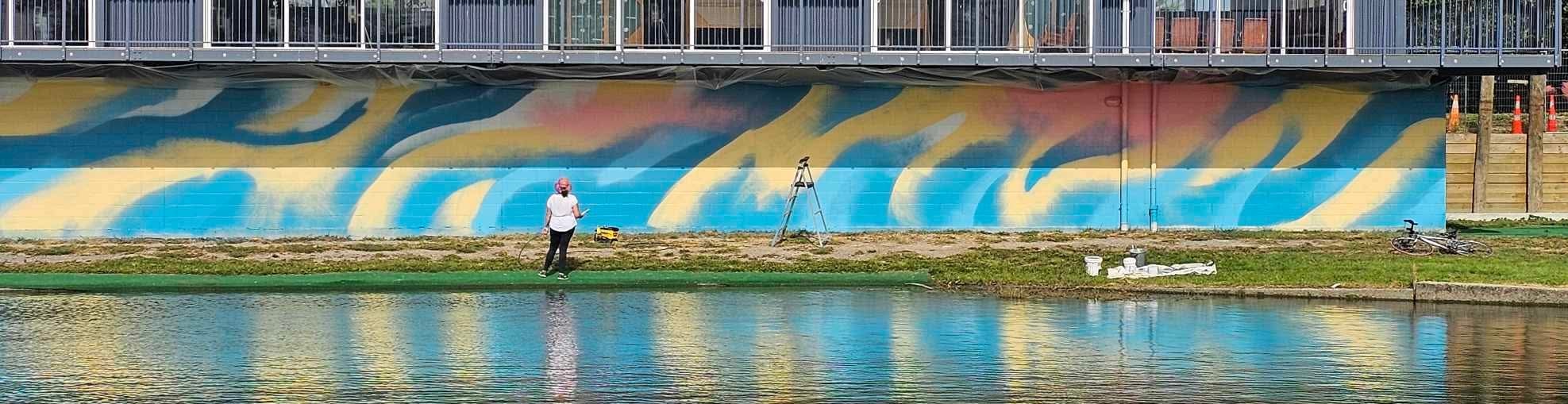 Mel walking along the riverbank in front of the finished Pulling Together mural at Cure Boat Club