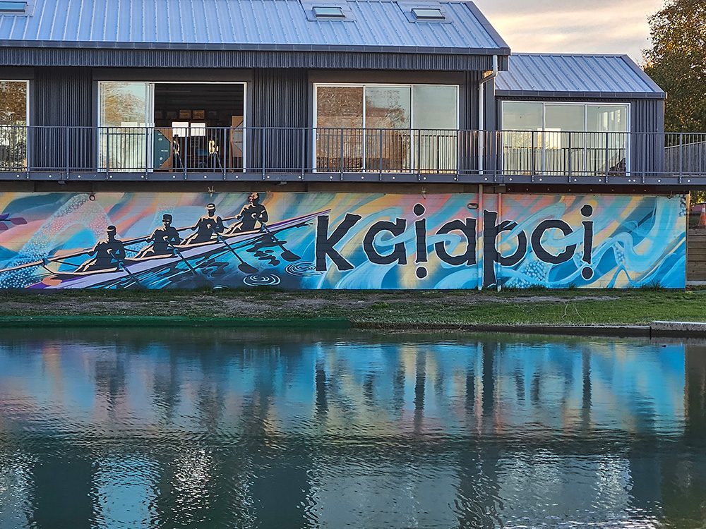 Pulling Together mural — golden hour view of the clubrooms and 'Kaiapoi' lettering at Cure Boat Club