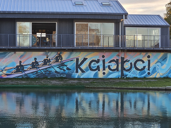 Pulling Together mural — rowers in a boat over a swirling water pattern with 'Kaiapoi' lettering, on Cure Boat Club's riverside wall