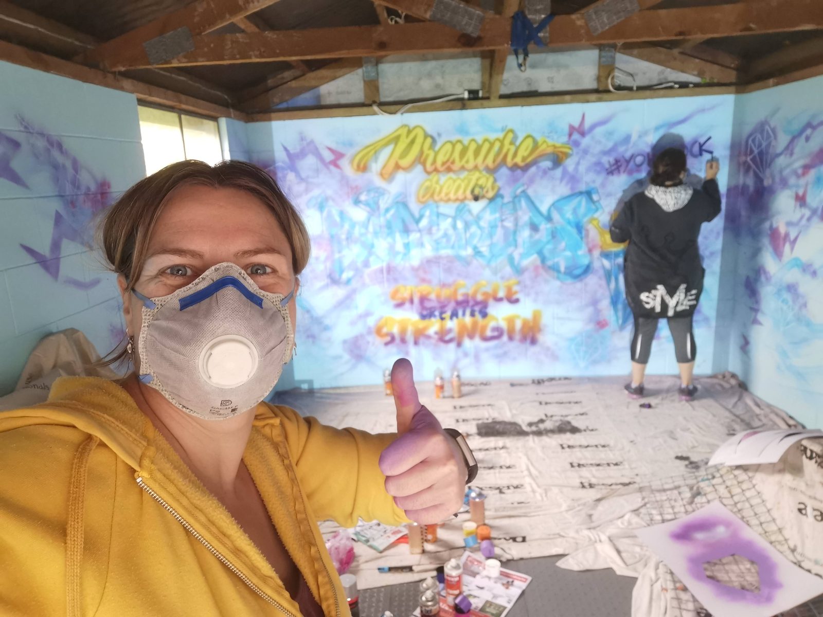 You Rock mural in progress — Mel in a respirator mask giving a thumbs up beside the unfinished design