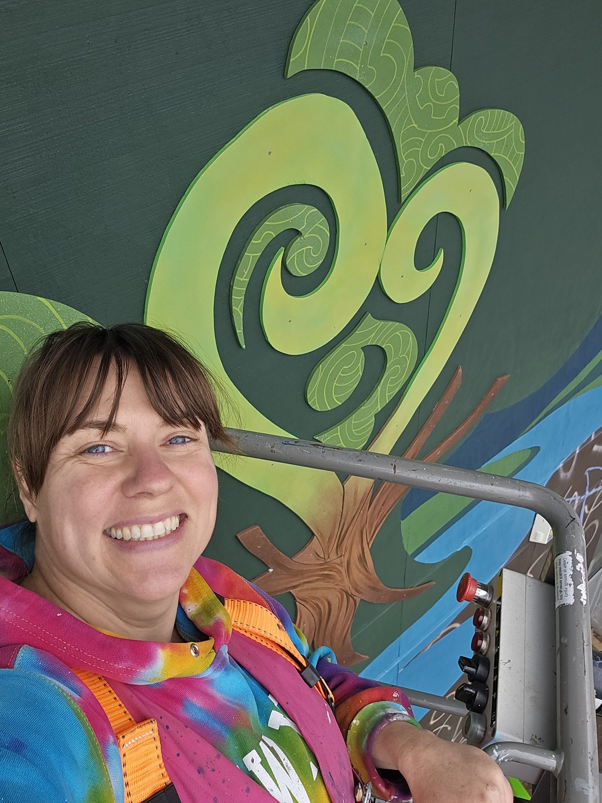 Around The Tree — Mel smiling beside the koru pattern with a painting tool in hand