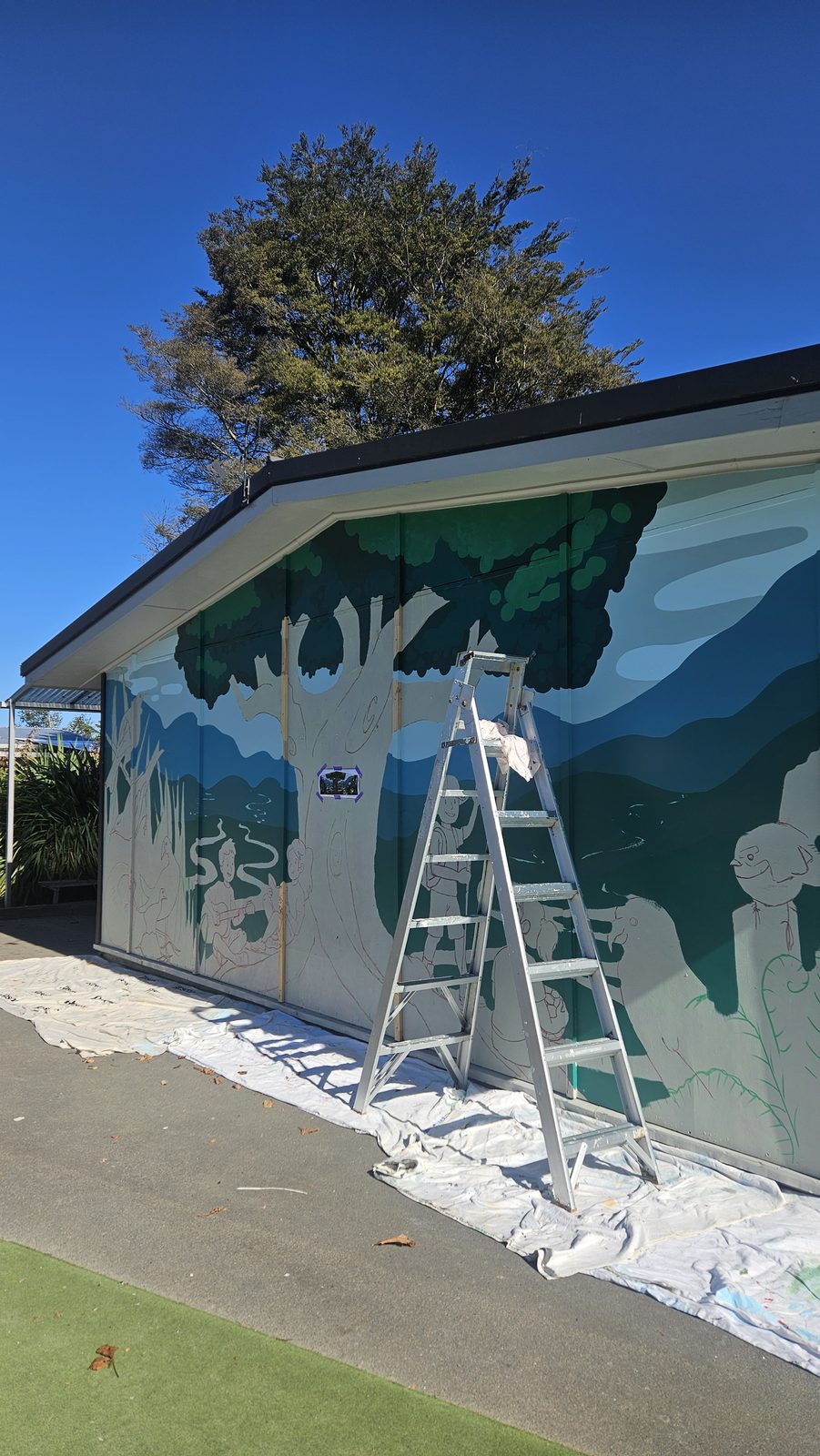 Flock Together mural in progress — blue-green hills blocked in with a ladder against the wall