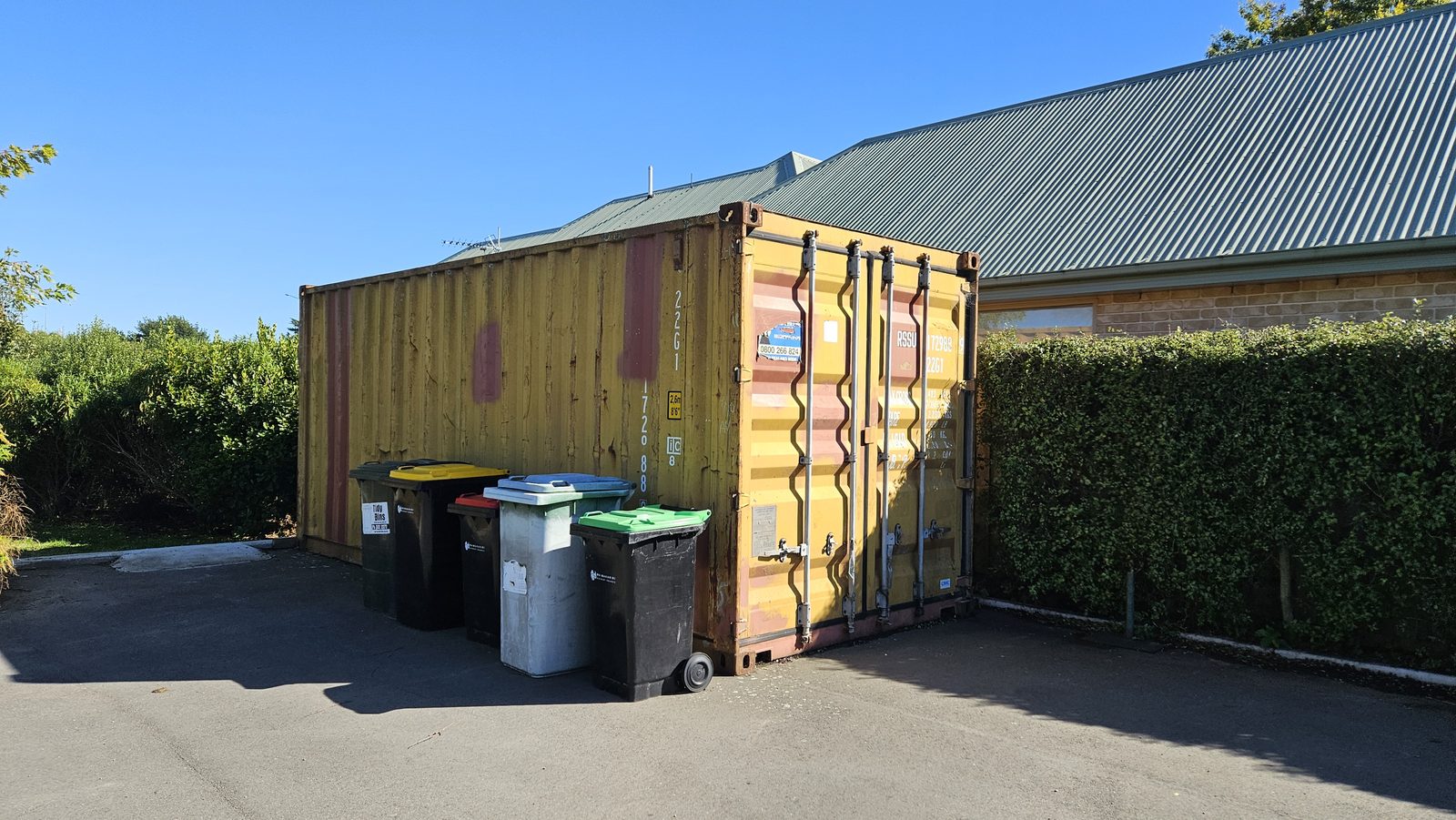 Growing Together — the worn shipping container before the mural, between hedges in the car park
