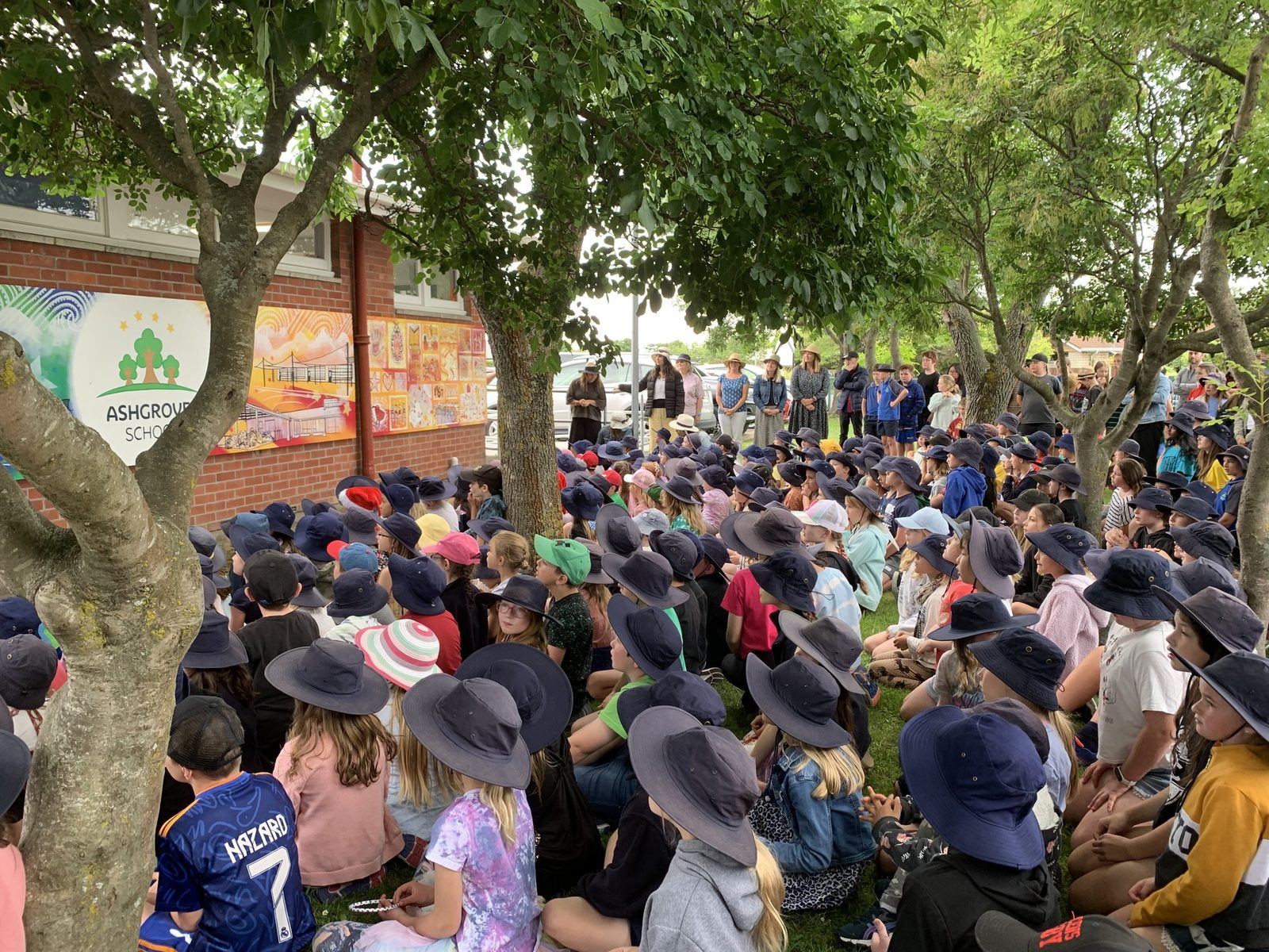 Our Stars — wide view of students seated under the trees for the mural unveiling