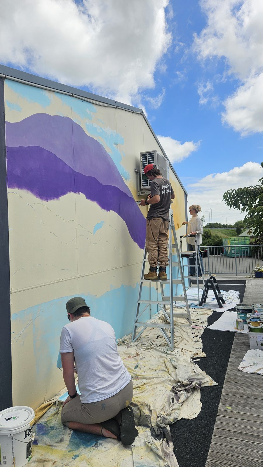 Turangawaewae mural in progress — purple mountains and green hills being blocked in on the wall