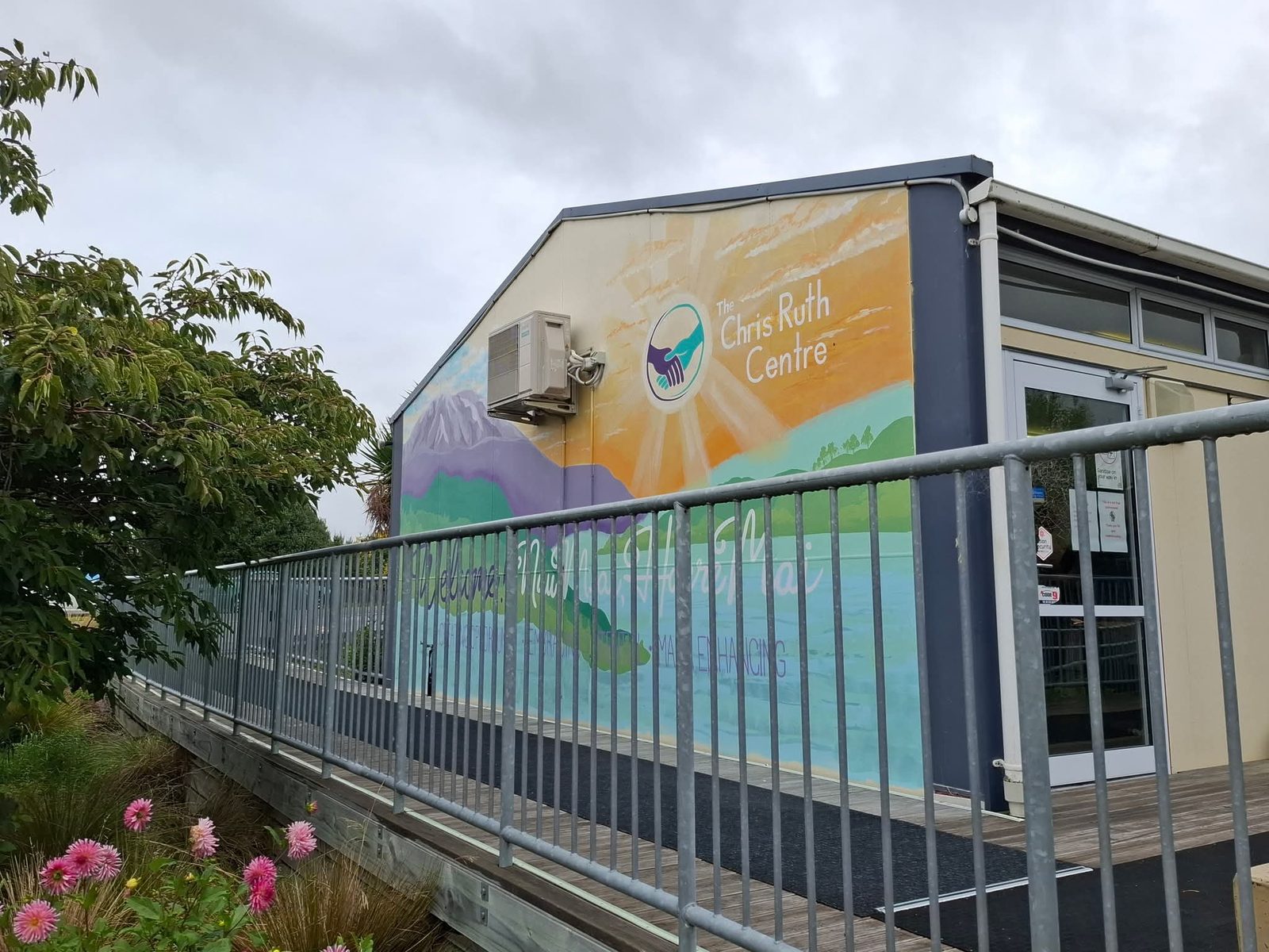 Turangawaewae mural — wide view of the building and mural from the footbridge, with flowers in the foreground