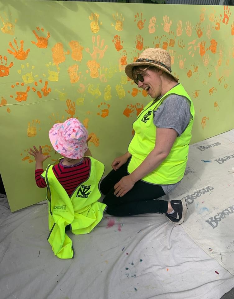 Weaving Together — Mel and a preschooler adding handprints to the mural together