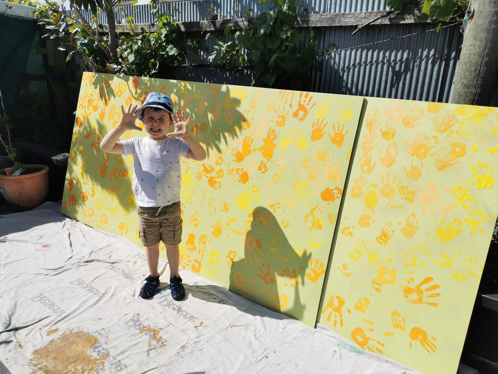 Weaving Together — a child standing beside a handprint panel in the sun