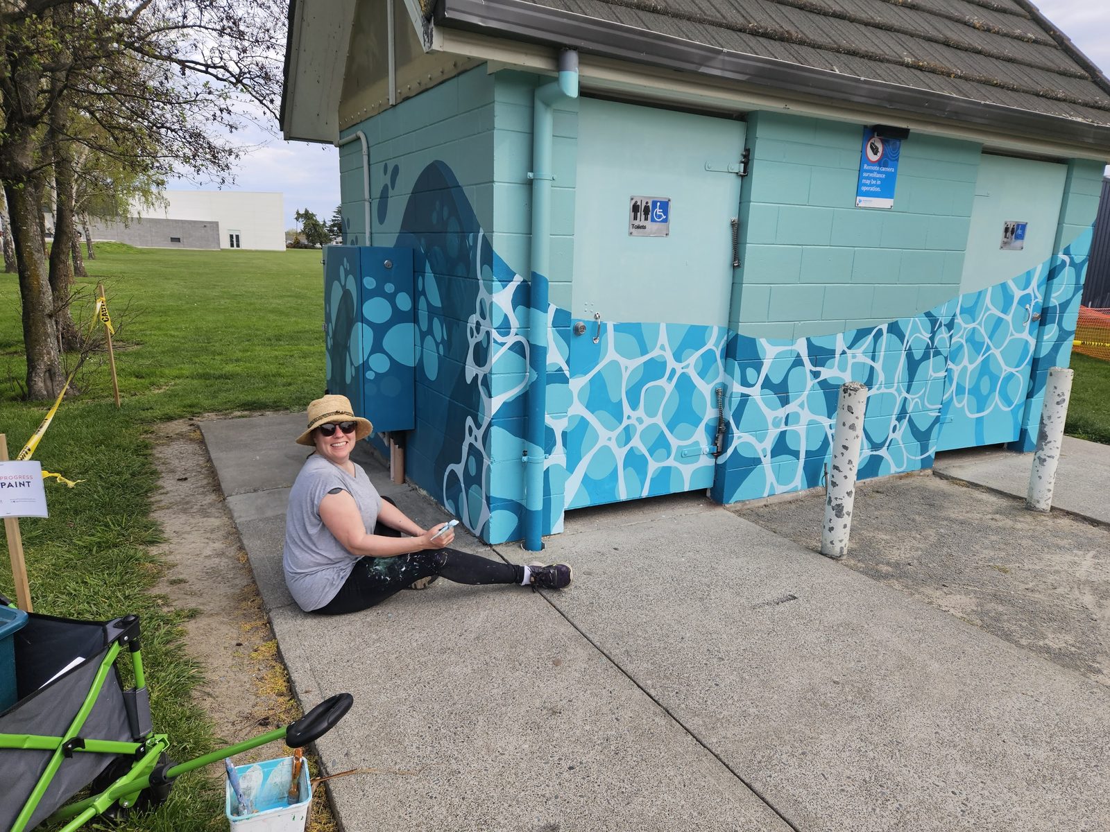 Flowing With Whio mural in progress — Mel sitting beside the building with painting supplies