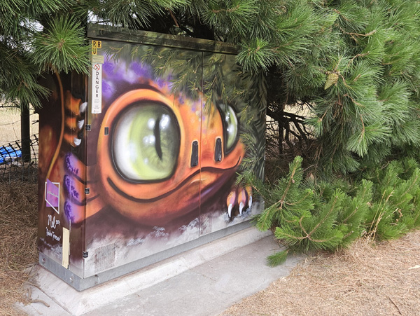 Hidden Treasure mural — a dragon's eye and face peeking out from a Chorus street cabinet tucked into pine trees