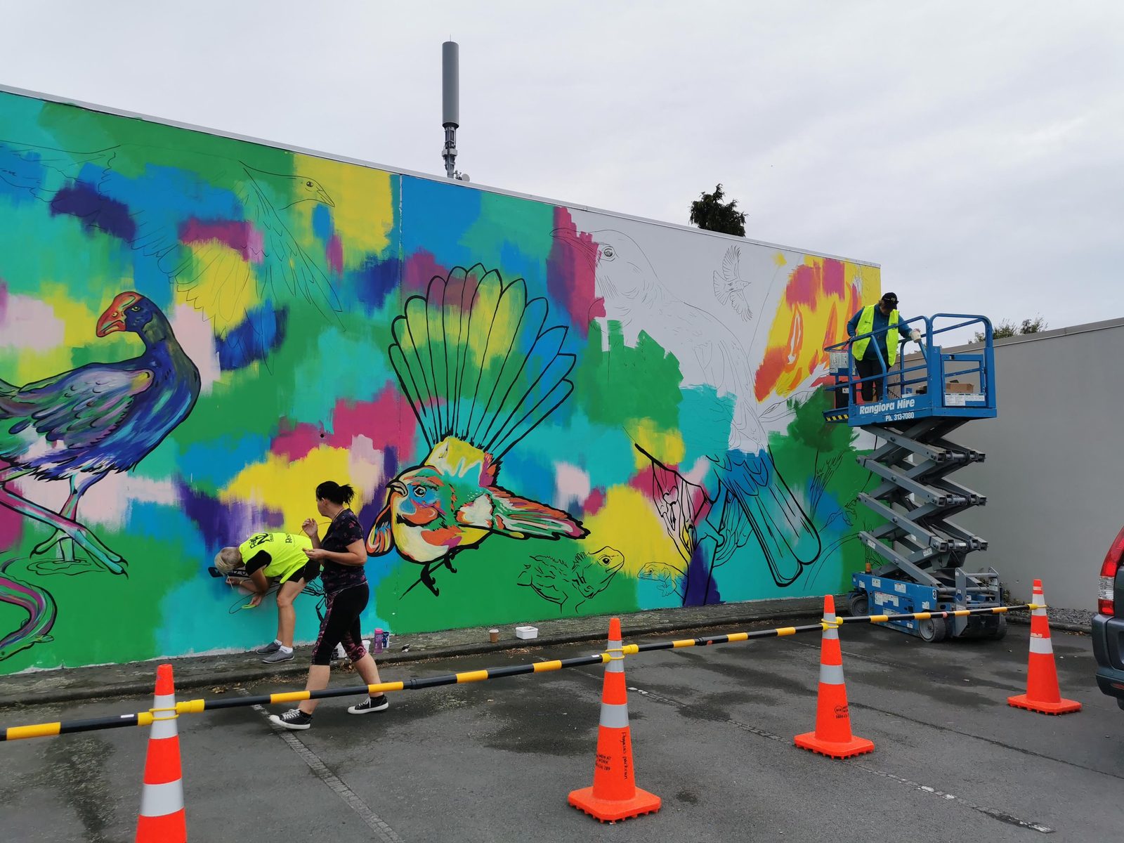 In Our Hands mural in progress — painters working on the base of the wall beside road cones