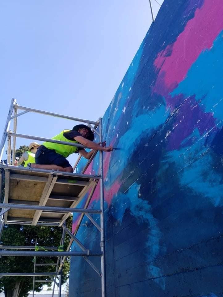 In Our Hands mural in progress — scaffolding against the carpark wall as the design takes shape