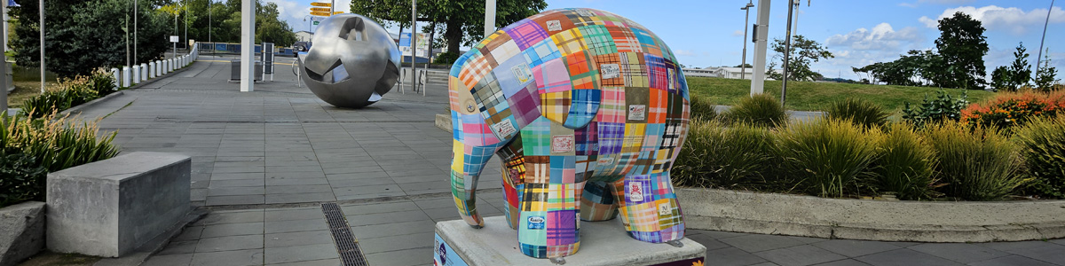 Mammoth Wolley sculpture — wool-blanket patchwork elephant sculpture on display in a public plaza, with a silver sphere sculpture in the background